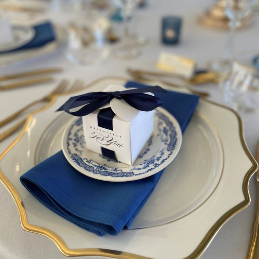 Dinner party place setting with gold rimmed white charger plate, blue floral side plate, gold cutlery, blue napkin, boxed gift
