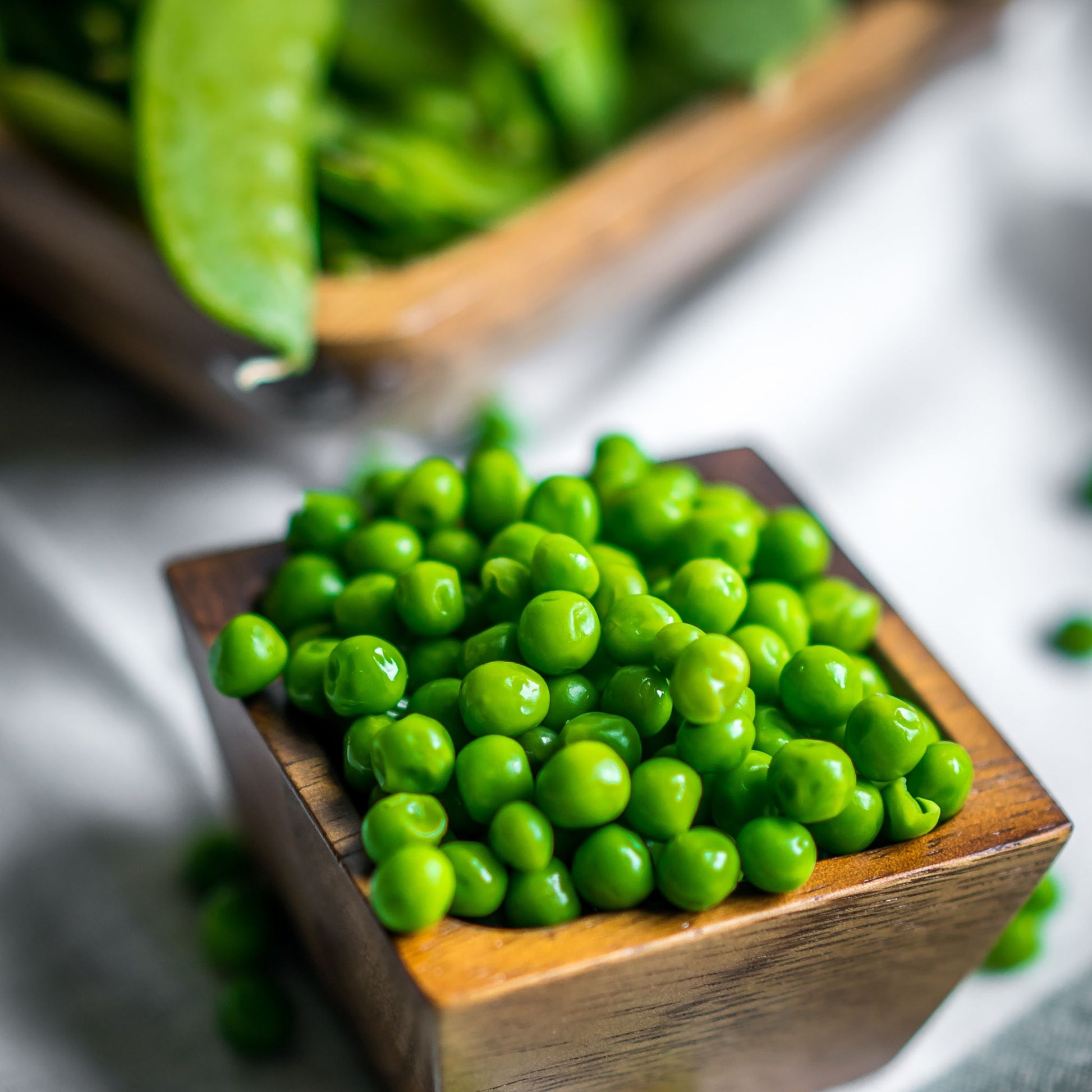 Wooden square bowl full of shelled green peas, pea pods in the distance