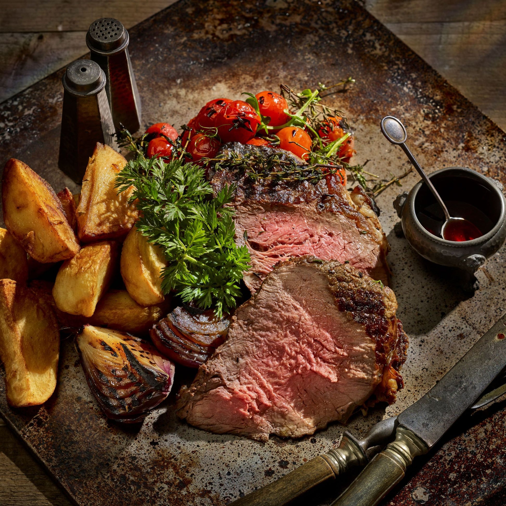 Sliced steak on a platter with potatoes, tomatoes, parsley and pot of sauce with carving knife and fork