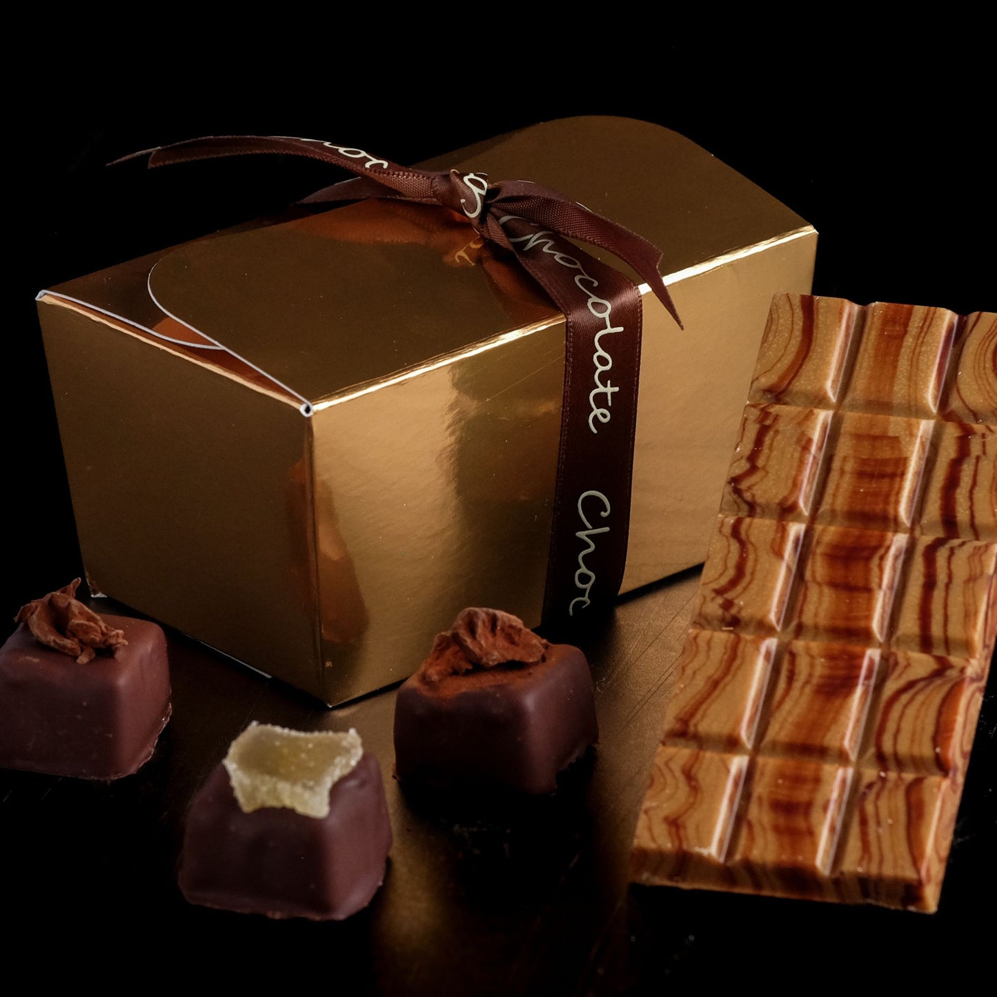 Gold chocolate box with a ribbon, surrounded by chocolate truffles and a bar of chocolate on a dark background.