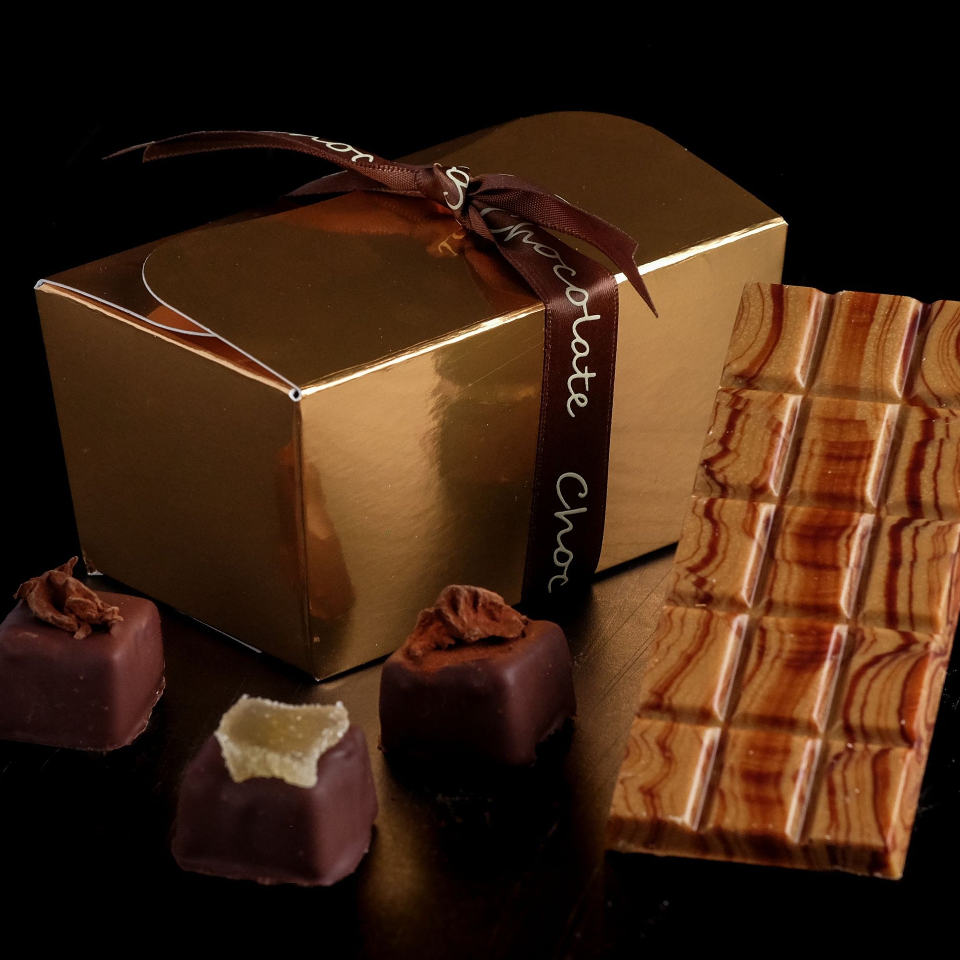 Gold chocolate box with a ribbon, surrounded by chocolate truffles and a bar of chocolate on a dark background.