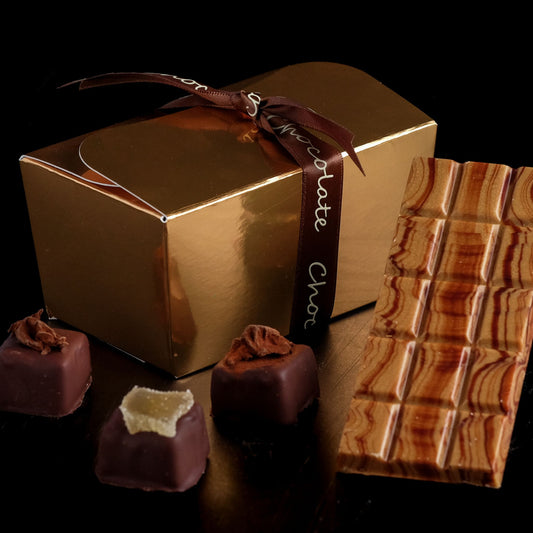 Gold chocolate box with a ribbon, surrounded by chocolate truffles and a bar of chocolate on a dark background.