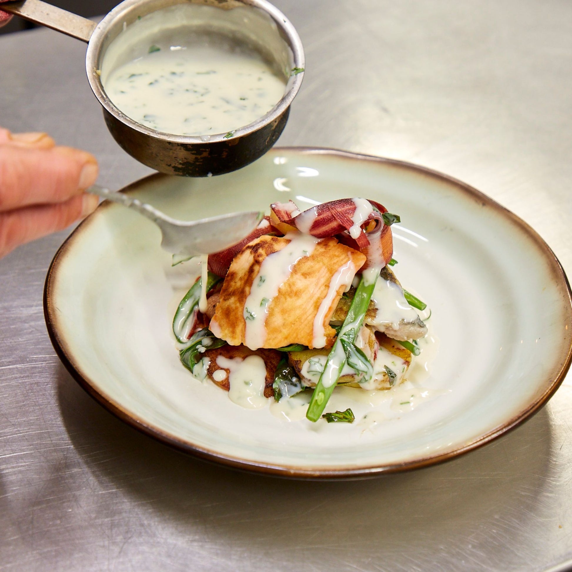 Round plate with cooked piece of fish on pile of green beans and vegetables man's hand pouring white sauce with a spoon from a small pan