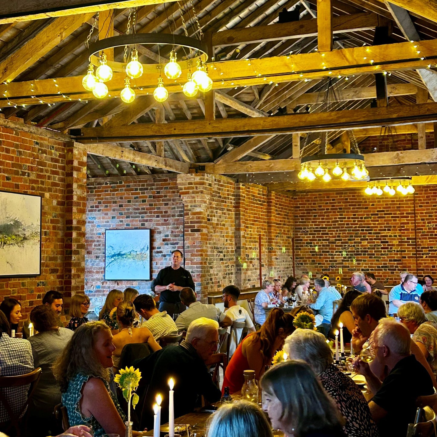 People sitting down at two rows of tables in a brick barn and a chef talking